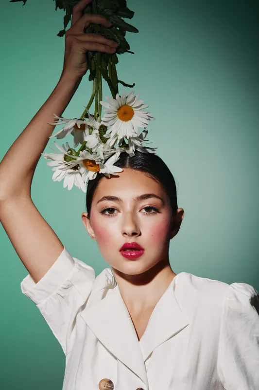 Young woman in a white shirt holds daisies above her head with elegant expression. Soft light, pastel background, beauty and fashion concept emphasizing natural aesthetic and creativity.