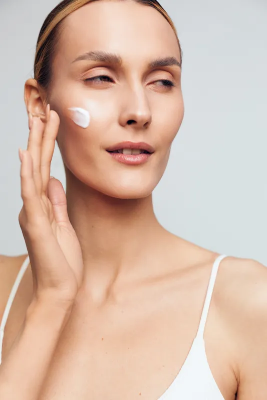 Young woman applies white cream to her cheek in a close-up shot, with soft studio lighting creating a calm, skincare focused mood.