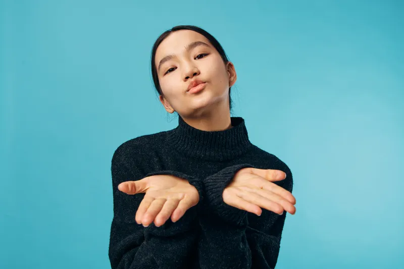Young woman blowing kiss with both hands in black sweater on blue background. Portrait of female with confident expression and casual style for positive gesture and emotion.