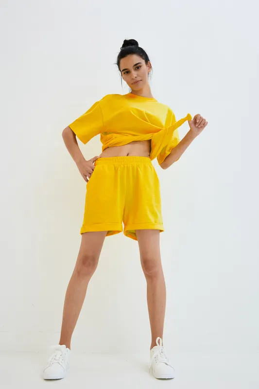 young woman in yellow casual outfit posing indoors against white wall, sporty loose T-shirt and shorts, confident stance, minimalistic bright background