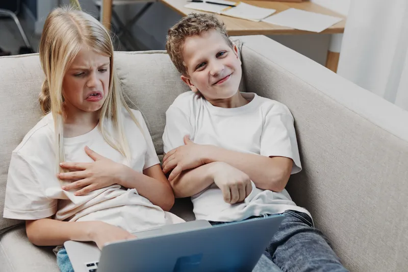Two children sitting on a beige couch using a laptop, girl showing disgust and boy smiling confidently. Concept of sibling contrast, emotions, technology use, and childhood moments.