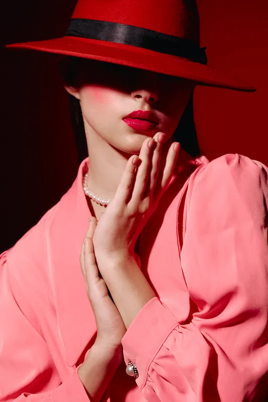Fashion portrait of a woman in red lipstick wearing a red hat with a black ribbon and a pink blouse. Her face is partially covered by the hat brim. Beauty and elegance concept.