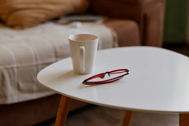 Cozy home scene with a white round table, a mug, a cup, and a pair of glasses resting beside it, warm natural light, soft colors, inviting relaxed atmosphere