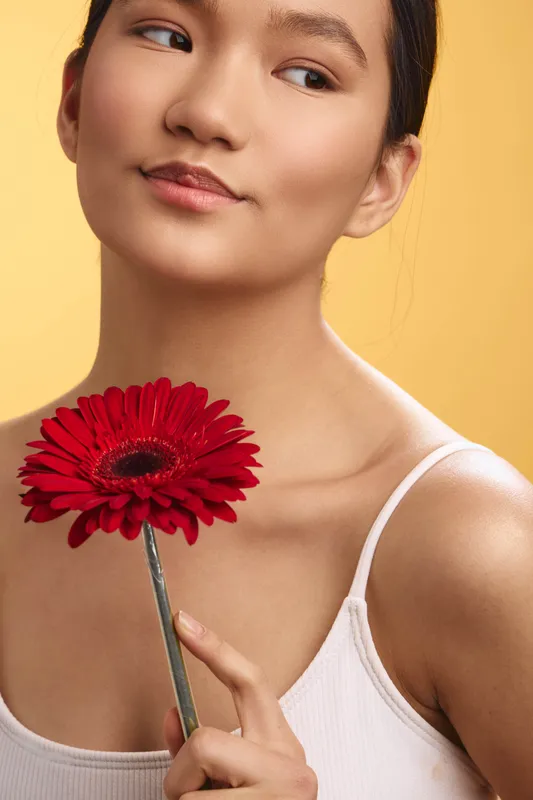 asian woman gently holds a bright red flower near her shoulder, calm expression and soft lighting create a serene portrait with natural beauty and quiet mood.