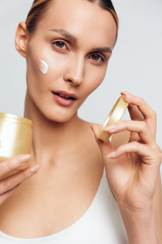 Young woman with clear skin applies face cream from a jar while looking at the camera in a bright studio setting with minimal makeup and natural expression.