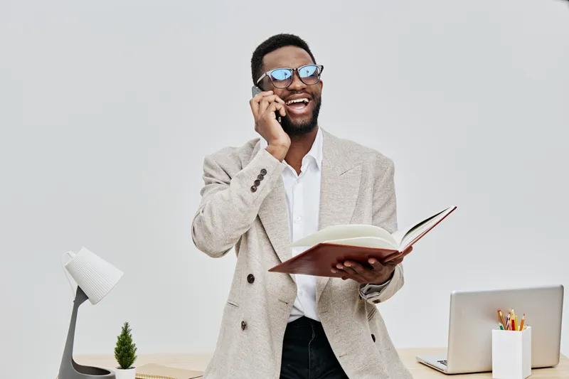 Smiling man talking on the phone while holding a notebook, dressed in a beige blazer and white shirt, fashion style, professional workspace, indoor office environment, cheerful mood, modern lifestyle 