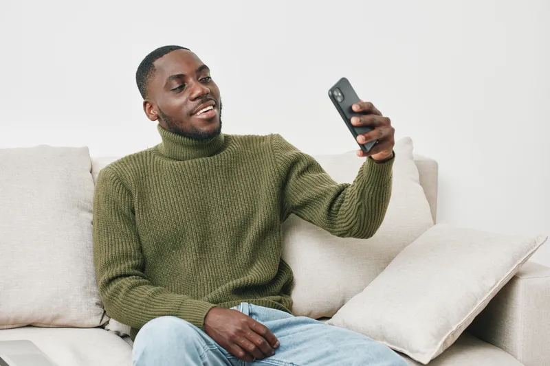 Smiling young Black man in a cozy green turtleneck sweater, enjoying a video call on his smartphone while sitting on a comfortable sofa in a bright living room