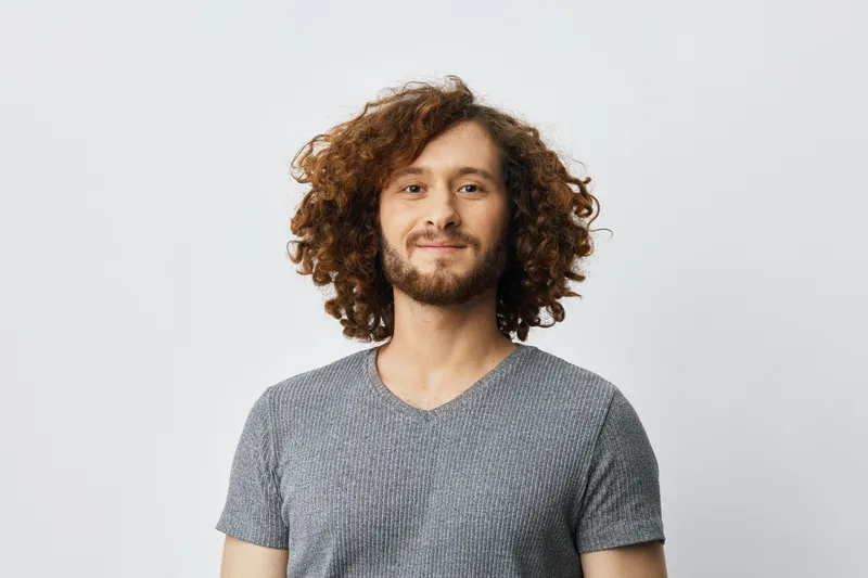 Young man with curly hair smiling against a light background in casual attire, showcasing confidence and warmth, ideal for lifestyle and beauty themes