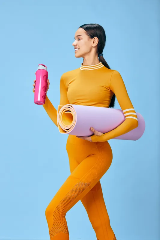 Active woman in bright orange athletic wear holding a pink water bottle and a rolled yoga mat against a blue studio background, showcasing fitness, exercise and lifestyle in a vibrant wellness image.