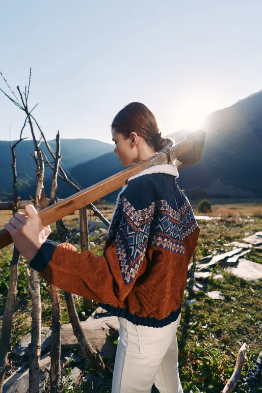 Woman with guitar outdoors in mountains at sunset, walking along trail wearing patterned sweater and light pants, backlit scene with natural landscape, peaceful travel and music.