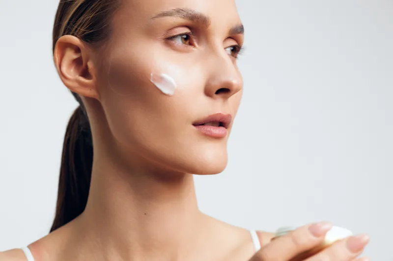 A young woman with clear skin applies cream to her cheek while holding a cosmetic jar. The minimal background highlights skincare and beauty care concepts.