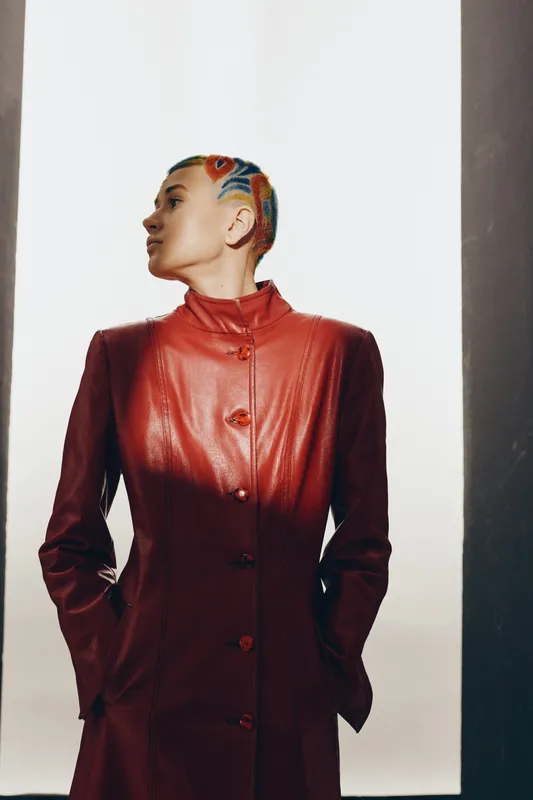 Woman with colorful braided hair wearing a red leather coat stands confidently indoors against a bright minimalist background, showcasing modern style and attitude.