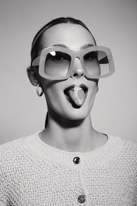 Young white woman with pale skin and slicked hair wears oversized square sunglasses and earrings, sticking out her tongue playfully in studio with soft lighting.