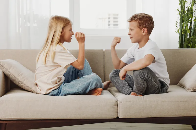 Two children sitting on a beige sofa facing each other, playing a hand game indoors in a bright living room with natural light and casual clothing. Concept of childhood, play, and friendship.