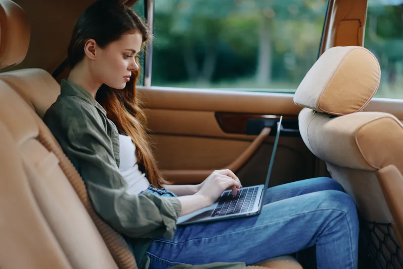 Young woman working on a laptop in the backseat of a car, showcasing a relaxed vibe and a modern lifestyle, ideal for remote work concepts.