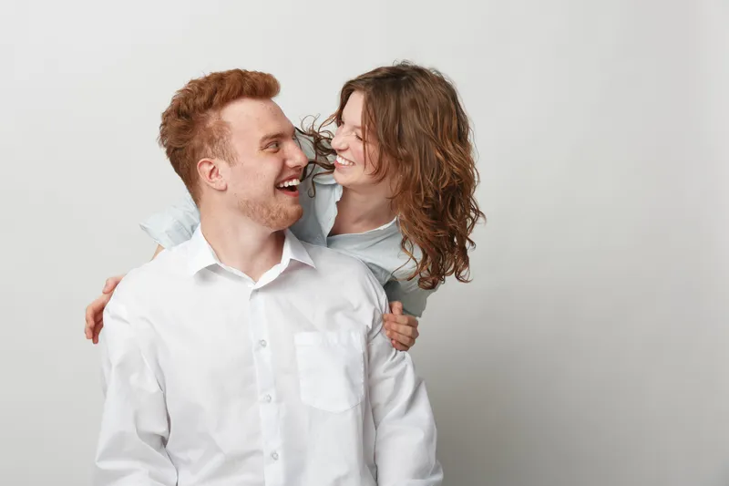 Happy young couple in white shirts laughing and embracing each other against a plain light background. Concept of love, relationship, joy, and togetherness in casual lifestyle.
