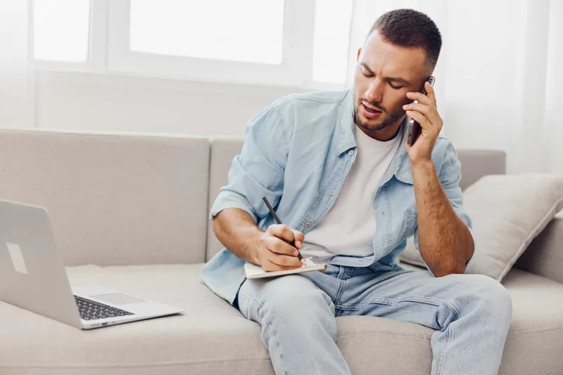 Focused man making notes while talking on the phone, dressed casually in a light denim jacket. The modern setting features ample natural light.