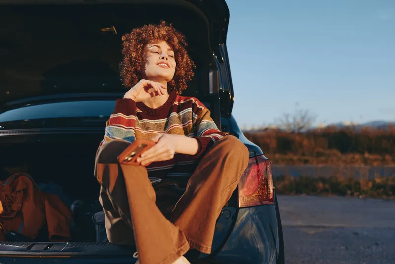 Woman lifestyle photo, relaxed in front of open car door outdoors, curly hair, cozy sweater and warm scarf, sunset on a rural road, calm travel mood, golden evening light, countryside horizon