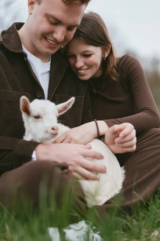 A couple outdoors gently cradle a white goat kid, sharing a warm moment in a bright rural setting. Lifestyle scene emphasizing companionship, nature, and simple joy.
