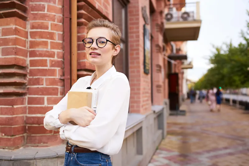 Женщина в очках с книгой в руках стоит на улице возле кирпичного здания учебного заведения