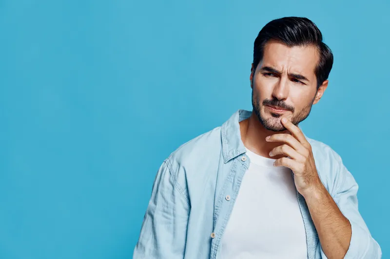 Curious thoughtful young man wearing light denim shirt poses against a solid blue background. He ponders, looking away while touching his chin thoughtfully.