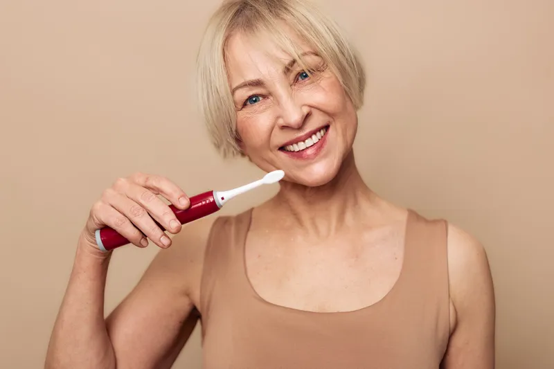 Smiling mature woman with short blonde hair cleaning teeth with electric toothbrush against beige background. Concept of oral hygiene, dental care, wellness, and daily routine.
