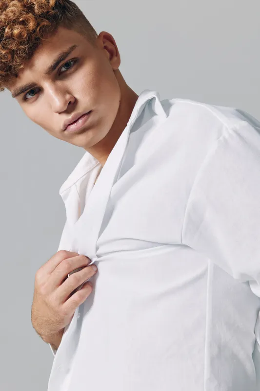Young man with curly hair wearing white shirt posing against gray background. Closeup portrait with serious expression and casual style in studio lighting.