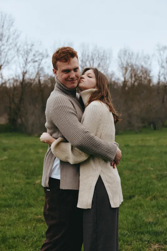 A couple hugs in a green park, wrapped in chunky sweaters under soft natural light. A warm, intimate moment that conveys romance, connection, and everyday tenderness in an outdoor lifestyle setting.