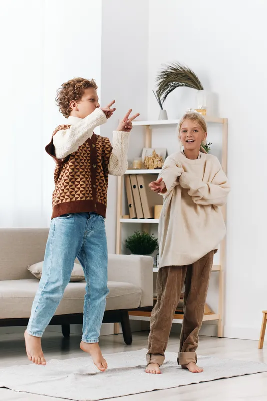 Two joyful children dance indoors, mid-movement, wearing cozy knits and jeans, in a bright living room with a soft rug, capturing a carefree playful moment.