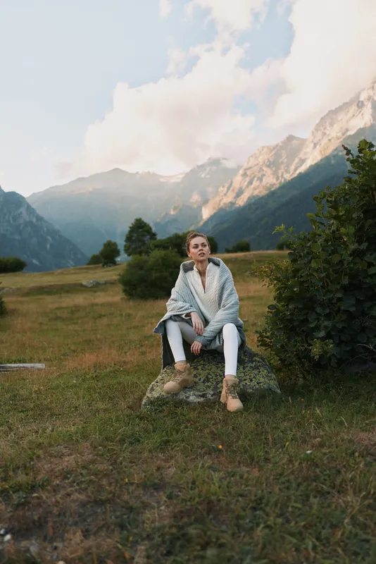 Woman lifestyle outdoors, seated in a meadow beneath mountains, relaxed pose, casual jacket and boots, soft light, natural colors, expansive alpine landscape behind