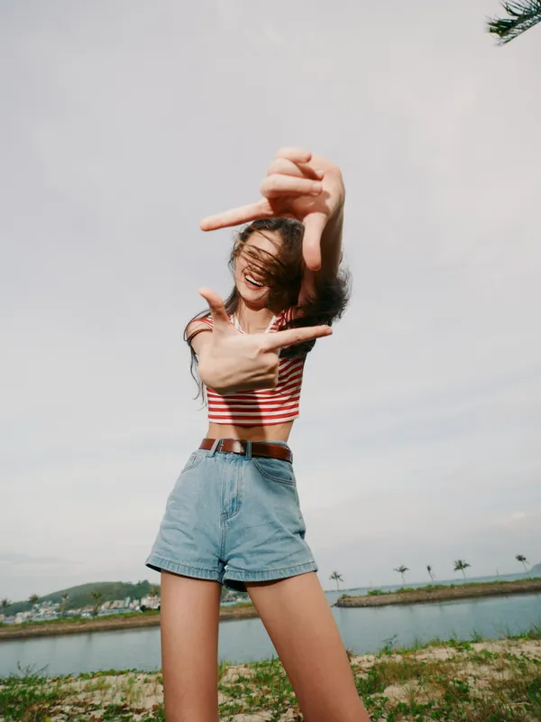 A young woman stands outdoors by a calm body of water, framing a shot with her hands. A cropped striped top and denim shorts convey carefree summer energy.