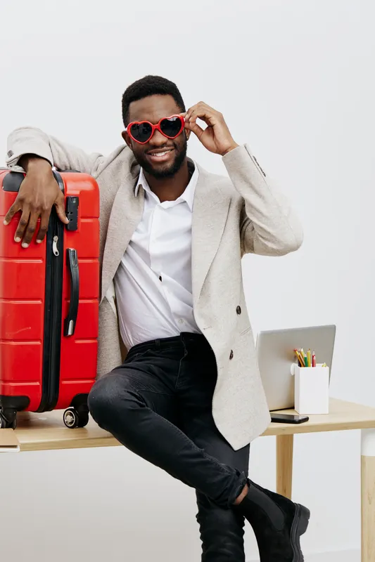Smiling man with sunglasses holding luggage suitcase and laptop in bright studio with minimal decor, joyful travel atmosphere, casual style, suggesting excitement and readiness for trip or vacation.