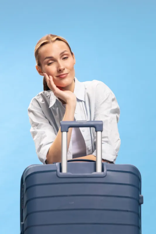 Tired young woman in casual white shirt leans on blue suitcase with closed eyes and relaxed expression. Concept of travel fatigue, waiting, and vacation preparation with blue background.