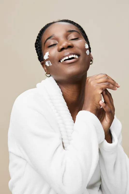Joyful black woman with skincare cream on her face, wearing a cozy white robe, smiling in a soft toned background, emphasizing beauty and self care