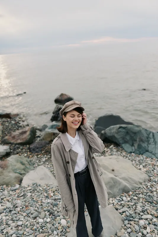 Smiling woman in warm coat and hat stands on rocky beach near calm sea under cloudy sky. Casual autumn outfit, nature, and outdoor lifestyle concept with happy female model.