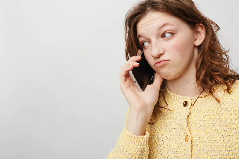 Young woman in yellow sweater talks on smartphone with skeptical expression, looking sideways. Concept of communication, doubt, and casual lifestyle in modern technology use.