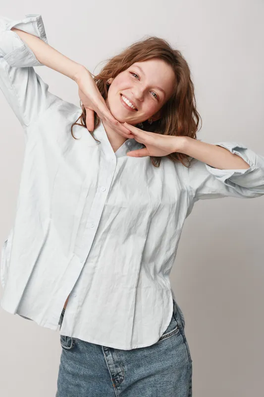 Smiling young woman in white shirt and blue jeans poses with hands under chin against light background. Casual style, happiness, youth, and positive mood concept in studio portrait.