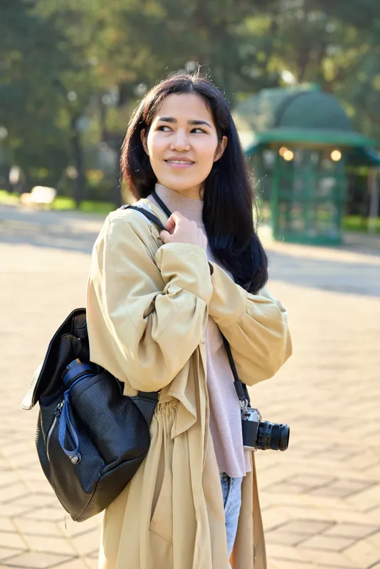 Traveler with camera and backpack in a sunlit park, casual coat and scarf, ready for street photography and urban exploration under warm light, curious expression.