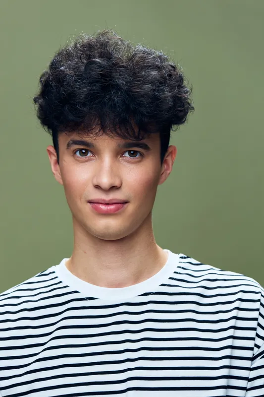 young man with curly dark hair wearing striped shirt standing against olive green background, looking confident and relaxed with direct eye contact