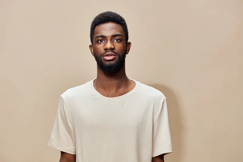 A young Black man with a confident expression wearing a light beige T shirt against a solid light brown background, emphasizing modern fashion and self assurance