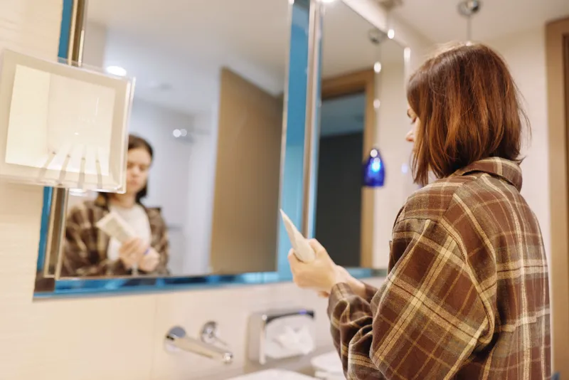 Woman wearing a plaid shirt stands at a bathroom vanity, looking into a mirror while holding a card, with warm lighting and soft reflection creating a calm, everyday moment.
