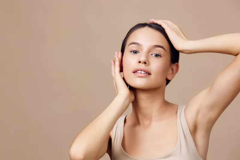 Young woman with a natural look posing against a beige background, showcasing healthy skin and a serene expression, emphasizing beauty and self care