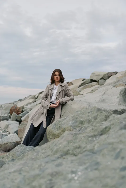 Young woman in long coat stands against large rocks under cloudy sky. Outdoor fashion portrait with natural background, casual style, and moody atmosphere.