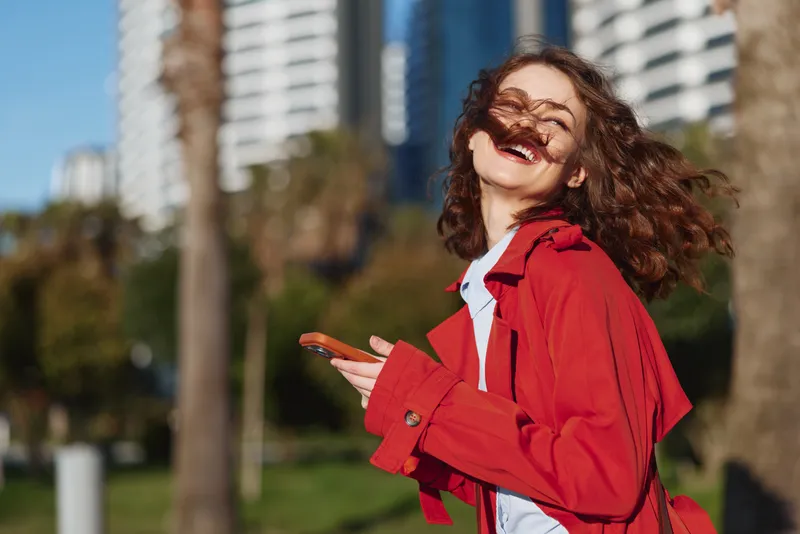 A joyful young woman in a red jacket enjoys a sunny day in an urban park, hair blown by the breeze, capturing a carefree moment of spontaneous happiness.