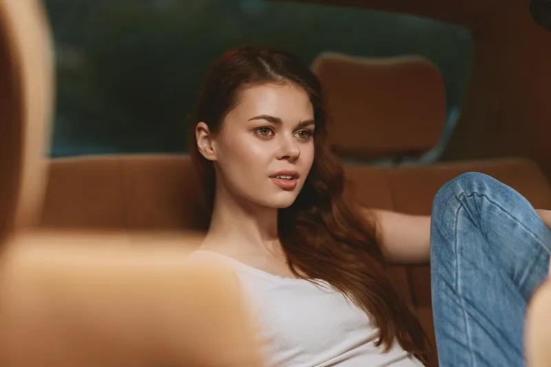 Young woman with long hair sits in a car's back seat, wearing a casual outfit, exuding a thoughtful expression in a cozy, relaxed setting.
