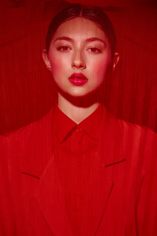 Young woman in red clothing and vibrant makeup poses behind red translucent fabric in a creative beauty and fashion portrait. Concept of high fashion and artistic style.