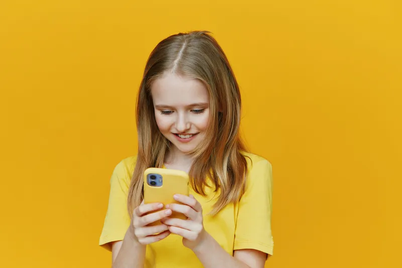 Young girl in a yellow shirt smiling while using a smartphone against a bright yellow background, expressing joy and modern lifestyle