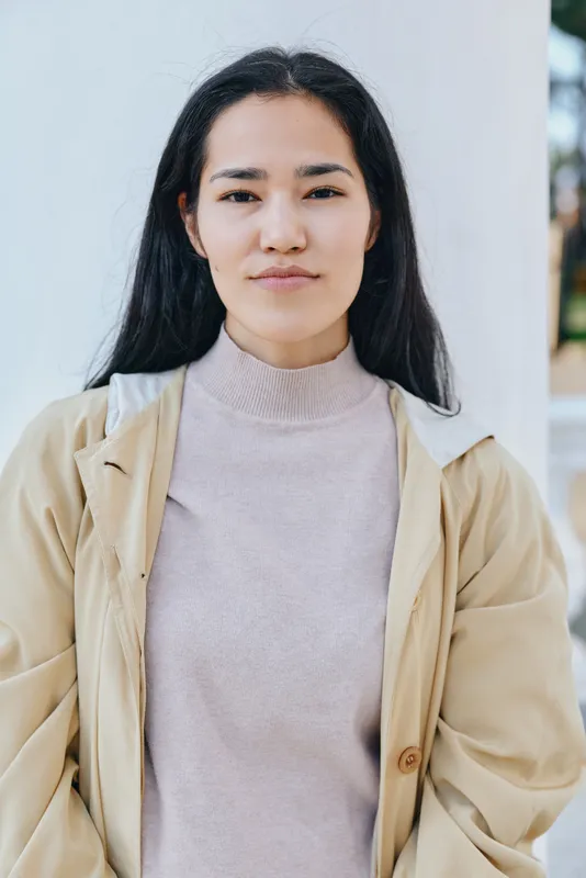 Woman in a light beige coat and soft pink sweater stands outdoors by a pale wall, calm expression, casual street style, natural light, contemporary fashion, everyday wear, relaxed pose