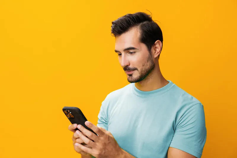 Мужчина на желтом фоне, улыбается, показывает на телефон, в очках
