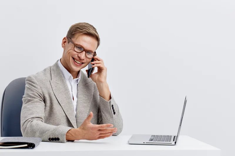 smiling man talking smartphone casual wear glasses sitting white table laptop remote work digital nomad communication concept